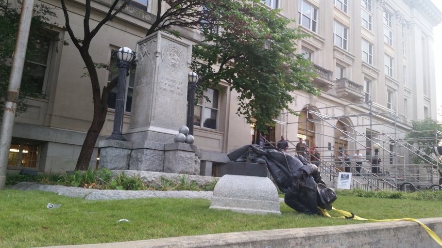 The fallen monument lays crumpled on the grass with the stone base in the background. Law enforcement officers survey the scene. 