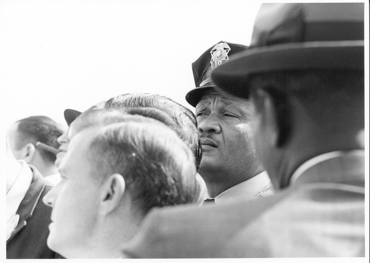 A group of men at the Wallace-for-president rally with the camera focused on the face of an African-American police officer. 