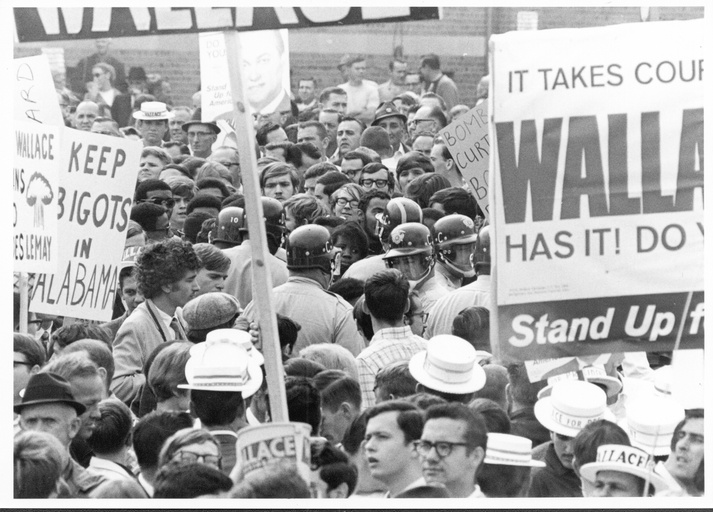 A large crowd of people with signs. Some police officers in riot gear visible. 