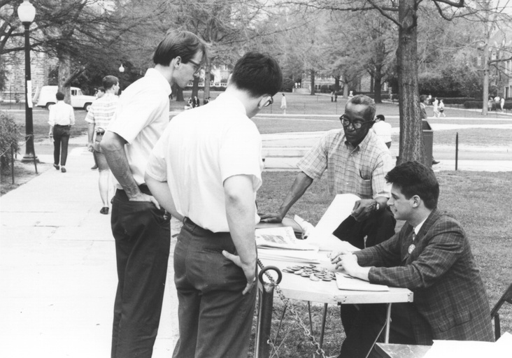 Two white men stand at an information table. A white male student and Oliver Harvey are behind the table.