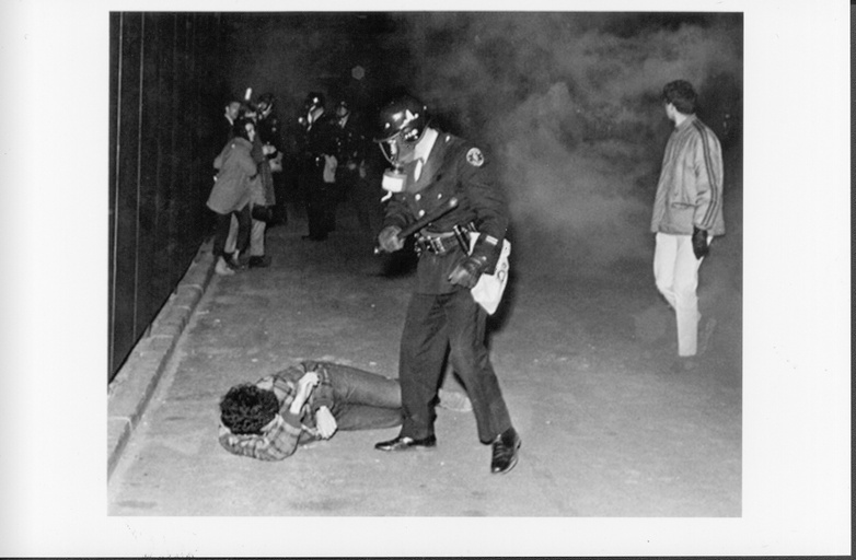 A police office holding a baton stands over a person laying on the ground. Other people and smoke are visible in the background. 