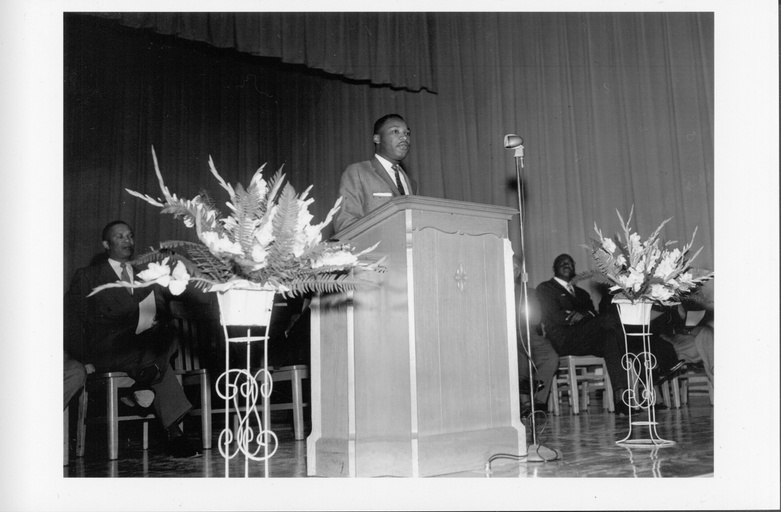 Dr. Martin Luther King stands behind a podium. Two other black men can be seen seated behind him.