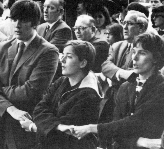 A group of white people stand with arms crossed, holding hands and appear to be singing. 