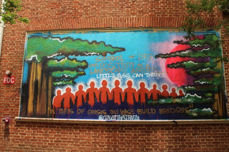 The profiles of a row of people holding hands painted in red with trees on either side of them and the words "symbol of life and possibility in landscape where little else can thrive" and "in times of crisis the wise build bridges."